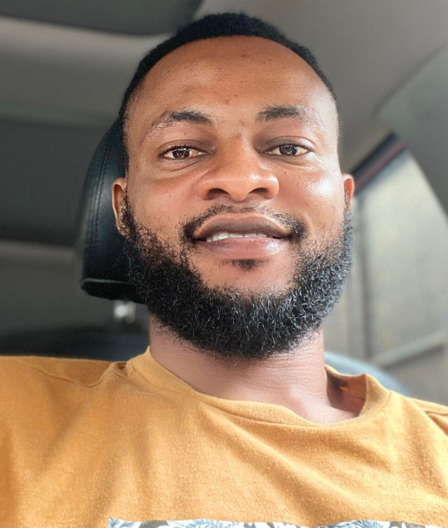 Portrait of Olumide Ibrahim Adesanya, a man with short hair and a full beard, wearing a yellow shirt. He is sitting inside a car and smiling at the camera.